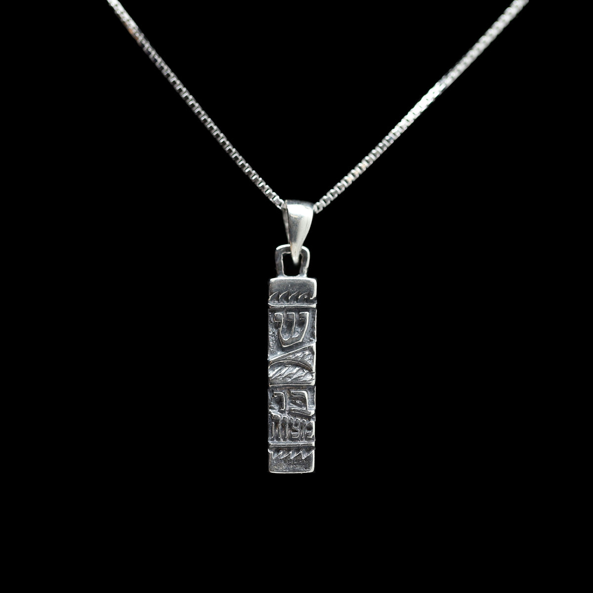 A silver pendant necklace featuring a vertically oriented rectangular bar engraved with stylized symbols as well as a hebrew letter shin and the words bar mitzvah in relief at the bottom. The pendant is suspended from a delicate silver chain against a solid black background.
