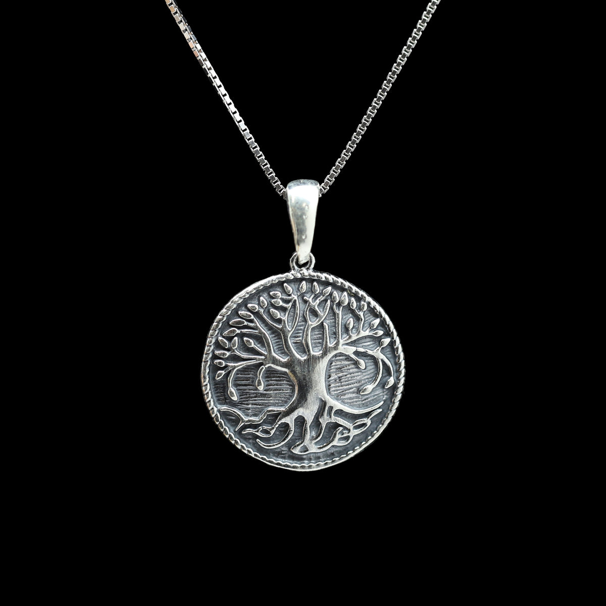 A silver pendant necklace featuring an embossed tree of life design on a round medallion, suspended from a silver box chain against a solid black background.