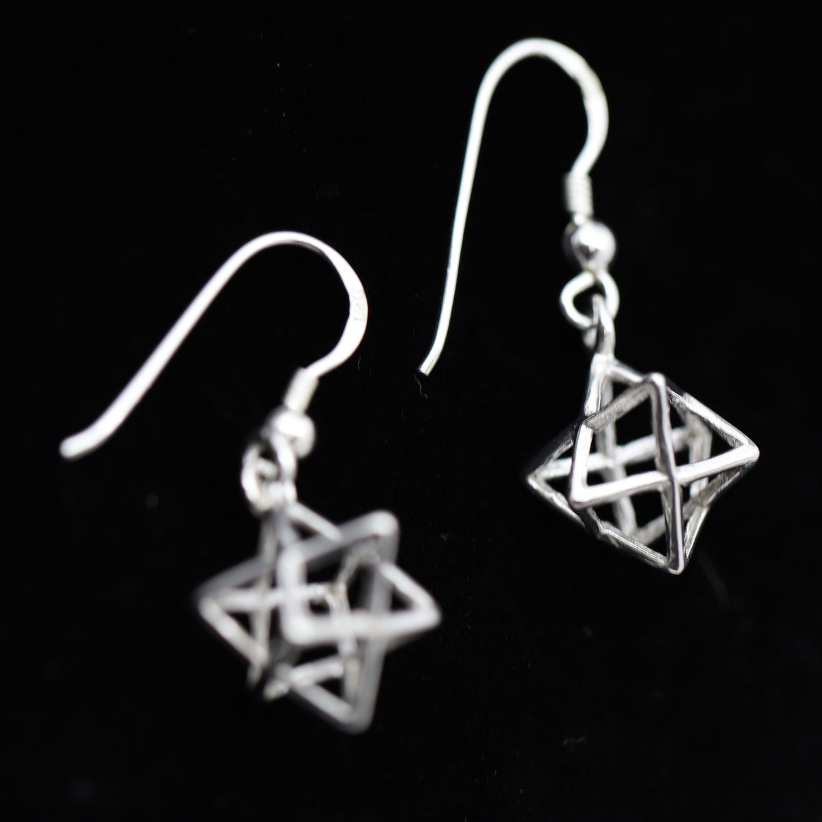 A pair of silver dangle earrings featuring interlocking geometric star and cube-like wireframe charms on fishhook wires, shown against a solid black background.